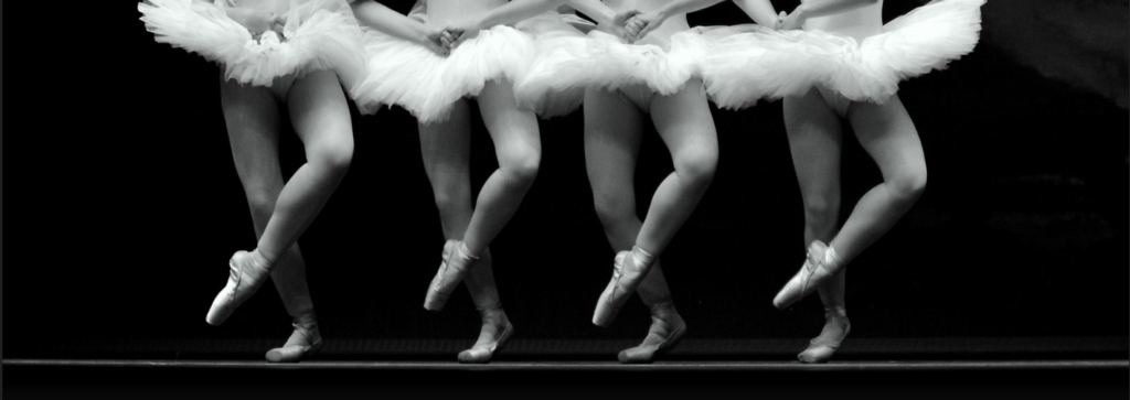 A row of four ballet dancers en pointe, one leg bent and slightly raised in front of the other.

